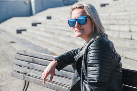 The woman sits on a wooden bench in jeans, a leather jacket, sunglasses outdoorsの写真素材
