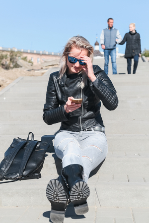 The woman sits on a stone ladder having extended legs in jeans, a leather jacket, sunglasses outdoors. To the utmostの写真素材