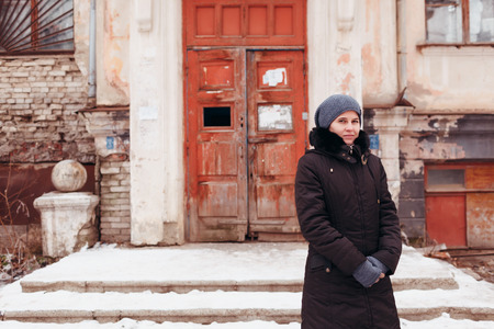 The woman stands near the old house in a black jacket in the winter