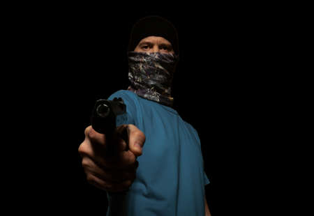 A criminal man in a mask on his face, aiming a gun at a camera, on a black background, isolated