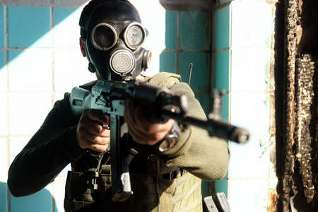 A military man in a black gas mask aims to point his automatic weapon at the camera