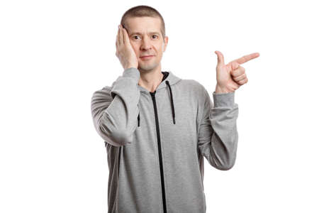 Surprised enthusiastic man in a hooded jacket, shows his index finger to the side, the palm of his hand at the head of the cheek. look into the camera. Isolated, on whiteの写真素材
