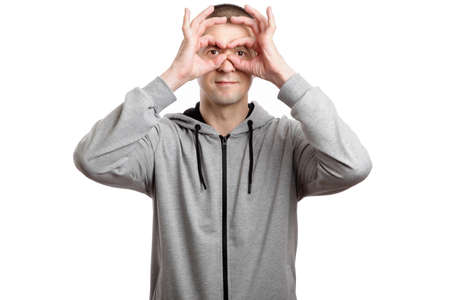 happy successful man, wearing a hooded jacket, shows the OK or okey sign, his hands like glasses in the eyes of his head. A look into the camera. Isolated, on whiteの写真素材