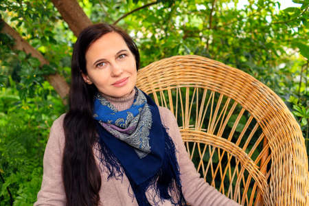 A woman in a blue headscarf and in a jacket sits on a braided chair in a garden among green plantsの写真素材
