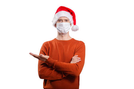 an isolated shot of a happy man in a medical mask from the coronavirus, in casual clothes, an orange sweater, a Christmas hat. With his finger he points to the left, looks into the camera. On whiteの写真素材