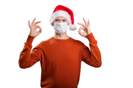 an isolated shot of a happy man in a medical mask from the coronavirus, in casual clothes, an orange sweater, a Christmas hat. Shows the gesture ok, looks at the camera. On whiteの写真素材