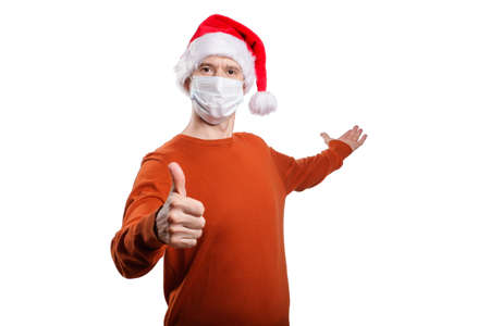 an isolated shot of a happy man in a medical mask from the coronavirus, in casual clothes, an orange sweater, a Christmas hat. gesture ok, the other hand points back, looks into the camera. On whiteの写真素材