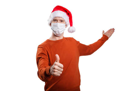 an isolated shot of a happy man in a medical mask from the coronavirus, in casual clothes, an orange sweater, a Christmas hat. gesture ok, the other hand points back, looks into the camera. On whiteの写真素材