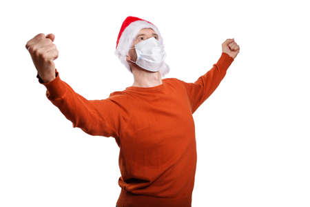 an isolated shot of a happy man in a medical mask, in casual clothes, an orange sweater, a Christmas hat. He clenched his fingers into a fist with happiness and luck, spread his hands to the sides. On whiteの写真素材