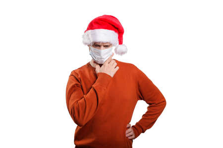 an isolated shot of an insecure man in a medical mask, in casual clothes, an orange sweater, a Christmas hat. He brought his hands to his head, looked down and made a decision, looked down. On whiteの写真素材