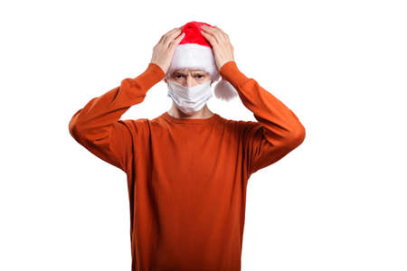 an isolated shot of an upset man in a medical mask, in casual clothes, a Christmas hat cap. He keeps his hands near his head from frustration, disappointment, trouble. On whiteの写真素材