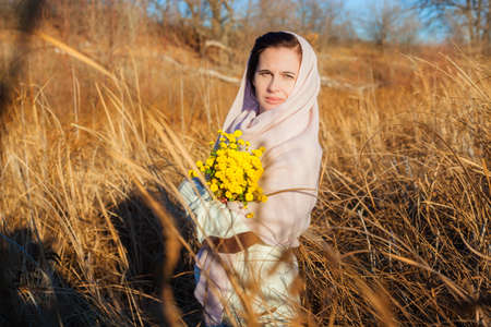 A serious girl in a field in the fall holds yellow wildflowers, looks into the camera. Dressed in casual clothes, sweater, scarf, oneの写真素材