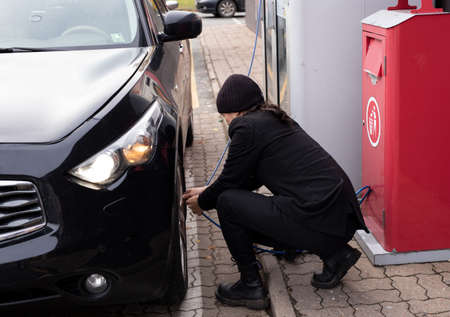 Young handsome adult man in black clothes standing on gas station and checking tire pressure.