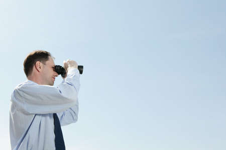 Businessman with binoculars against blue skyの写真素材