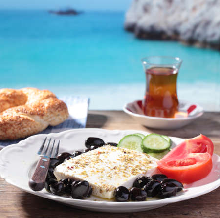 Breakfast with feta cheese, olives, simit, vegetables and tea against Mediterranean seaの写真素材