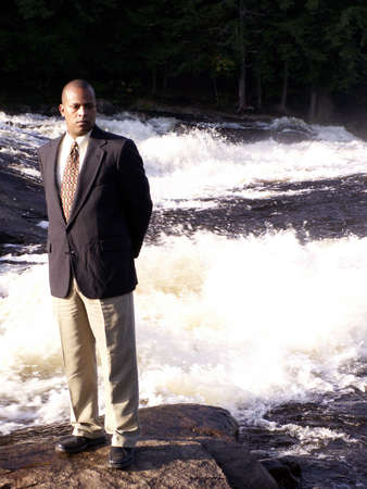 business man in a suit standing on a rock in front of a rushing riverの写真素材