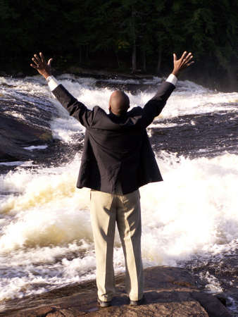 business man in a suit standing in front of a rushing river with hands raised in a triumphant gestureの写真素材