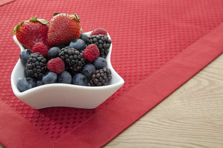 Fresh berries in an ornate white bowl on a red placemat.の写真素材