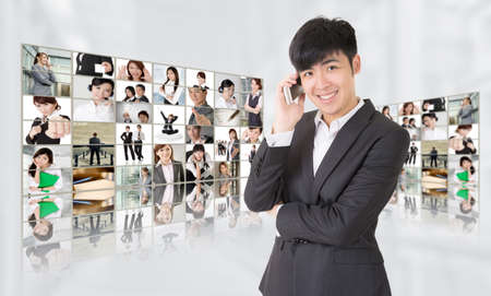 Young businessman talk on phone and stand in front of tv screen wall showing pictures of business concept by Asian business people. Concepts about business, communication, teamwork or social network.の写真素材