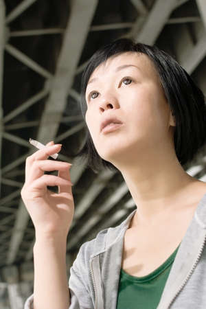 Closeup portrait of mature Asian woman smoking under bridge at city in daytime.の写真素材