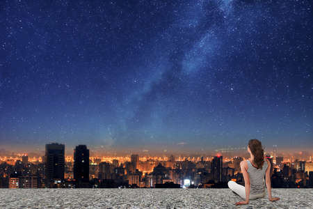 Asian woman sitting on roof, back to camera, and looking on the night city under starry sky.の写真素材
