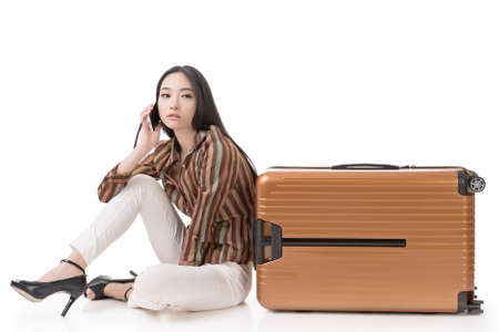Modern Asian woman sit on ground with a luggage and use a cellphone, full length portrait on white background.の写真素材
