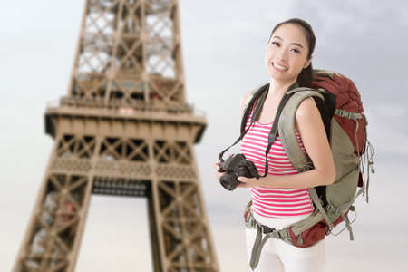 Smiling traveling Asian girl holding a camera and looking at you in front of famous Eiffel Tower in Paris, France.の写真素材