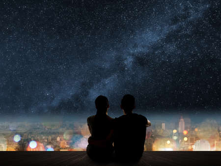 Silhouette of young Asian couple sit on wooden ground above the city under stars.の写真素材