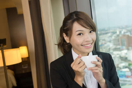 Asian business woman holding a cup of coffee and looking into distance near the window in hotel room.の写真素材