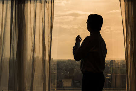 Silhouette of Asian businessman looking through the glass of window at sunset.