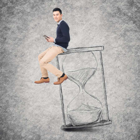 Asian young man sit on a hourglass and use tablet, concept of time management, technology, plan etc.の写真素材