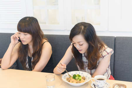 Asian woman eating lunch and her friend talk on cellphone in restaurant.