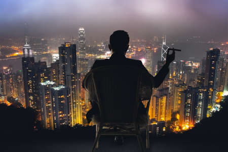Silhouette of businessman sit on chair and hold a cigar, looking the harbor of Victoria at Hong Kong, Asia.の写真素材