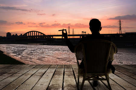Silhouette of businessman sit on chair and hold a cigar and looking at the city in night.の写真素材