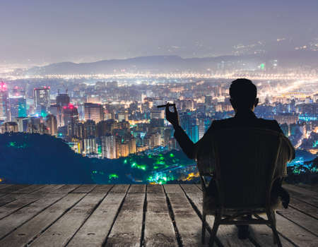 Silhouette of businessman sit on chair and hold a cigar and looking at the city in night.の写真素材