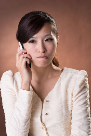 Angry Asian beauty holding cellphone, closeup portrait on studio brown background.の写真素材
