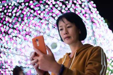 mature Asian woman using cellphone at night with lighting bulbの写真素材