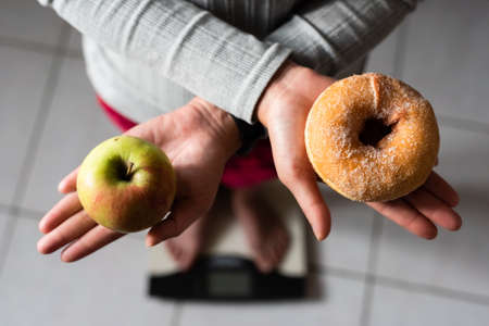 woman hold an apple and donuts and stand on scale at home, concept of healthyの写真素材