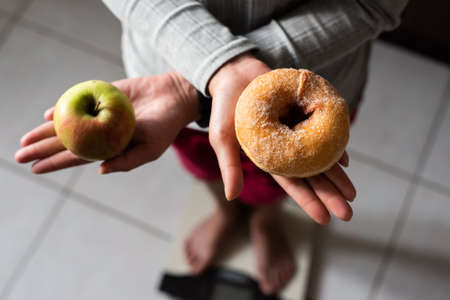woman hold an apple and donuts and stand on scale at home, concept of healthyの写真素材