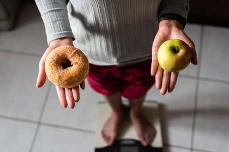 woman hold an apple and donuts and stand on scale at home, concept of healthy