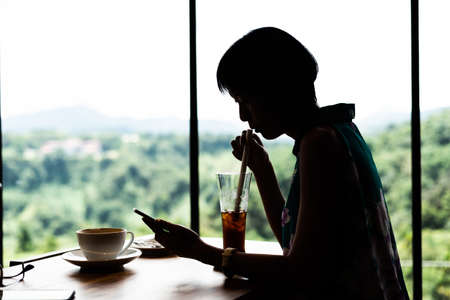 silhouette of Asian woman use mobile phone and get a cold drinks in the coffee shopの写真素材