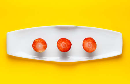 Three slices of fresh strawberry on a white elongated plate on yellow background. Horizontal image seen from above.の写真素材