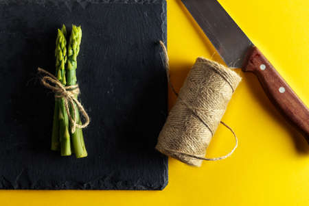 Asparagus. Bunch of fresh asparagus tied on a plate of slate with a knife and a roll of string on yellow background. Healthy vegetarian food. Horizontal image.の写真素材