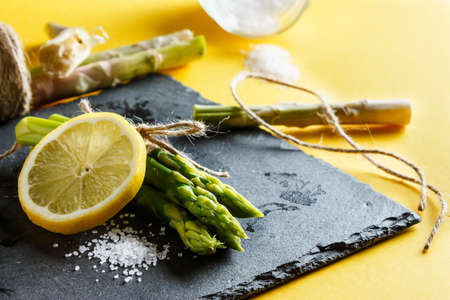Asparagus. Bunch of fresh green asparagus close up with coarse salt and a slice of lemon on a slate plate on yellow background. Rope pieces and esparraggos cut to the bottom. Healthy vegetarian food. Horizontal imageの写真素材