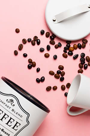 Coffee beans out of the coffee pot with the pot lid next to it and a white cup on a pink background.の写真素材
