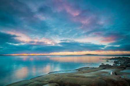 Blue, cloudy reflections over a calm ocean with a rocky coastline.の写真素材