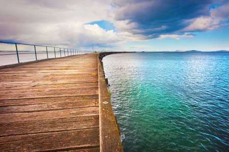 A long, curving, wooden jetty extends out over the ocean towards the distant horizon.の写真素材