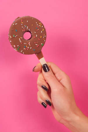 Women hand holds doughnut ice cream on a stick isolated on pink background, close upの写真素材