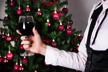 Elegant woman holding red wine glass in hand and decorated Christmas tree in background. Close up