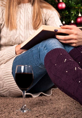 Young girl sitting and reading book and wine glass next to legs with warmers, Christmas tree in background, close upの写真素材